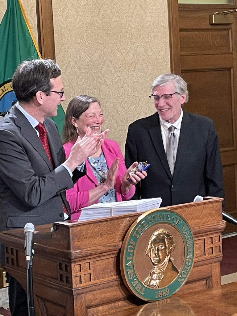 Washington Gov. Bob Ferguson, left, and state Sen. June Robinson applaud state Rep. Timm Ormsby, a veteran Spokane lawmaker who confirmed Wednesday that he will not seek reelection. Moments earlier, Ferguson signed the 2026 budget for which Ormsby and Robinson were the chief architects. (Photo by Jerry Cornfield/ Washington State Standard)