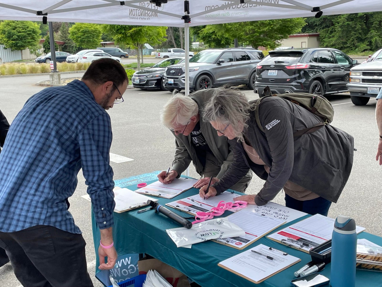 Josh Johnson (left) of Let’s Go Washington looks on as two more people sign petitions for a proposed ballot measure to repeal Washington’s new income tax on high earners. These signatures were gathered June 3, 2026 in Tumwater. (Photo by Jerry Cornfield / Washington State Standard)