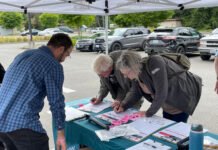 Josh Johnson (left) of Let’s Go Washington looks on as two more people sign petitions for a proposed ballot measure to repeal Washington’s new income tax on high earners. These signatures were gathered June 3, 2026 in Tumwater. (Photo by Jerry Cornfield / Washington State Standard)