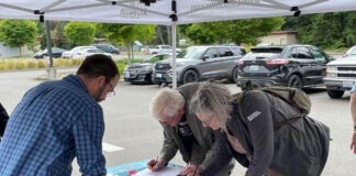 Josh Johnson (left) of Let’s Go Washington looks on as two more people sign petitions for a proposed ballot measure to repeal Washington’s new income tax on high earners. These signatures were gathered June 3, 2026 in Tumwater. (Photo by Jerry Cornfield / Washington State Standard)